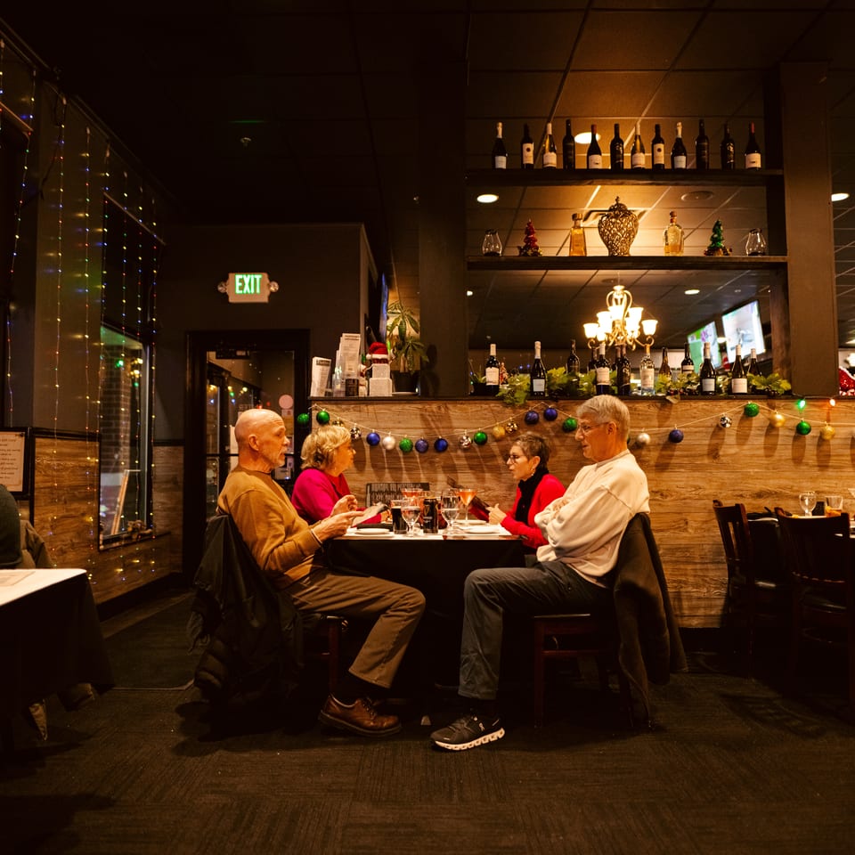 Warmly lit restaurant dining room with guests, drinks, wine displays, festive garlands, and colorful string lights.