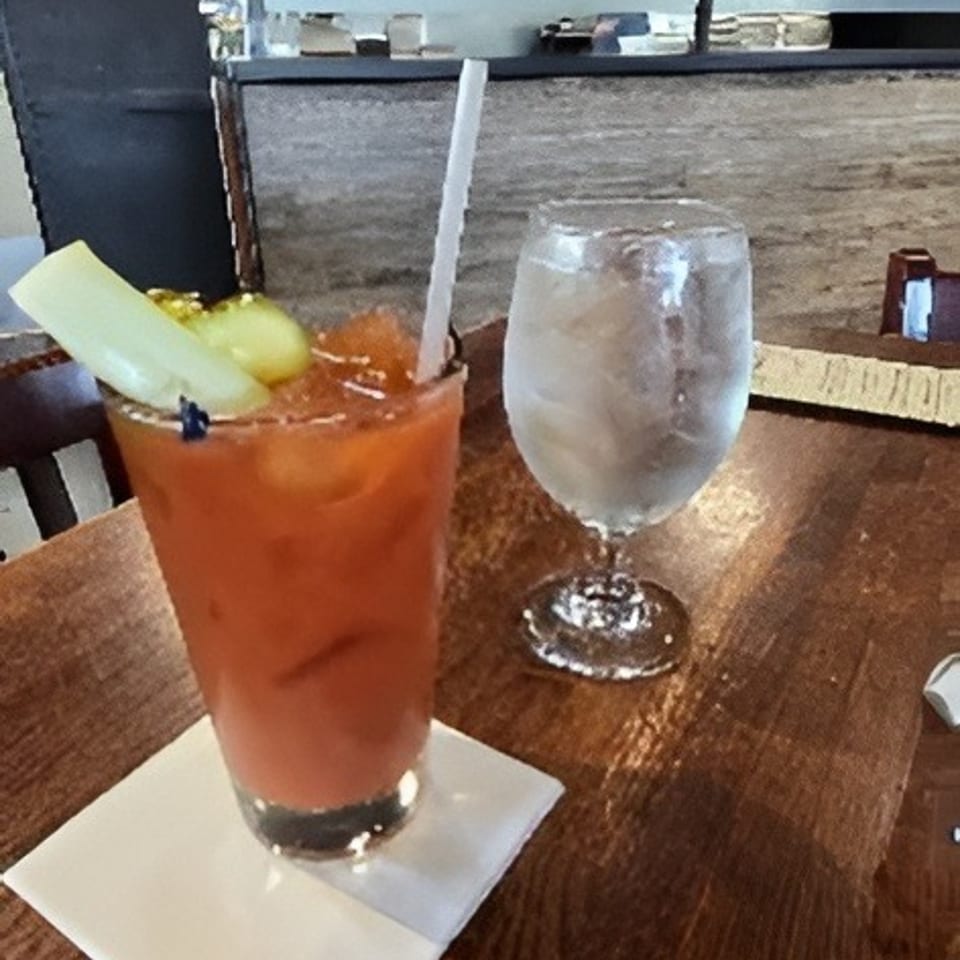 Bloody Mary garnished with celery and olives beside a glass of ice water on a wooden restaurant table.