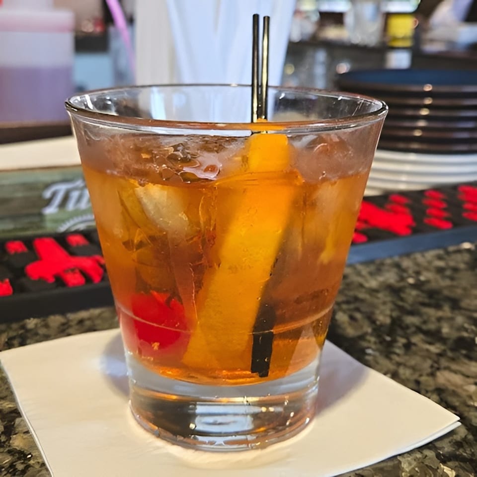 Amber cocktail with ice, orange peel, cherry, and black straw served in a rocks glass on a white napkin.