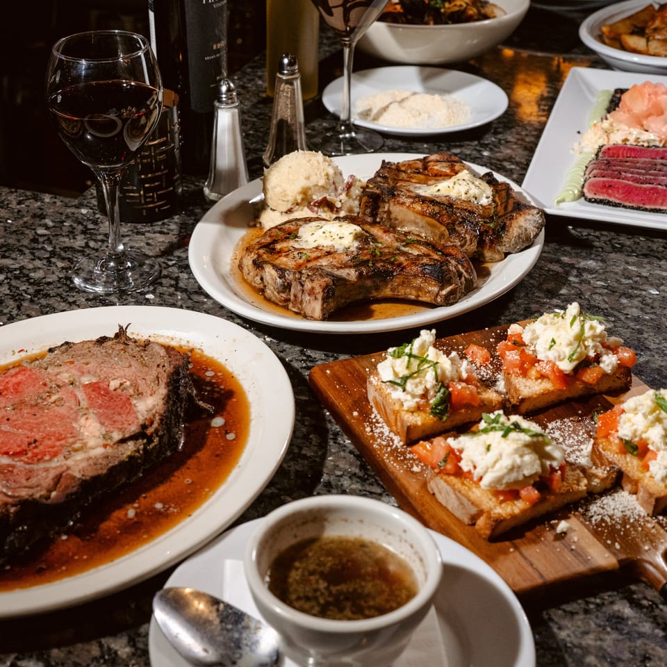 Restaurant table covered with grilled steaks, sliced roast beef, bruschetta, sushi, side dishes, and glasses of red wine.