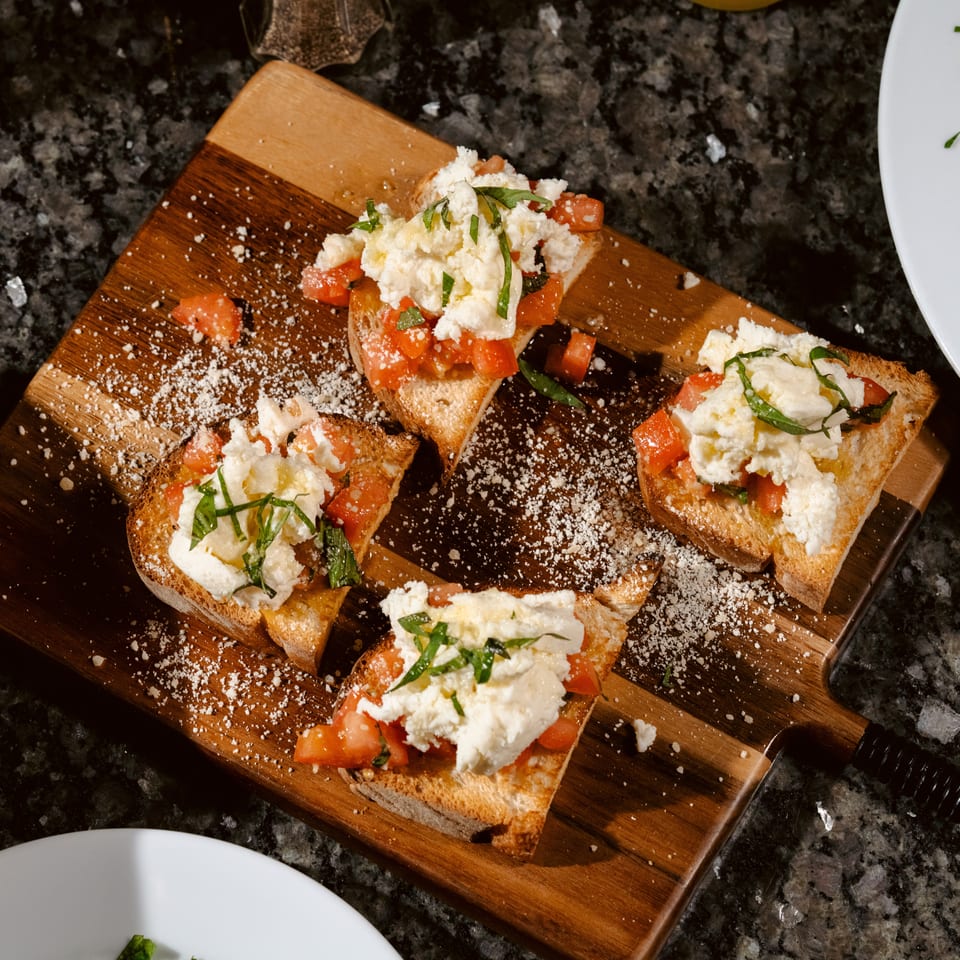 Toasted bruschetta topped with diced tomatoes, creamy cheese, basil, and grated cheese on a wooden serving board.