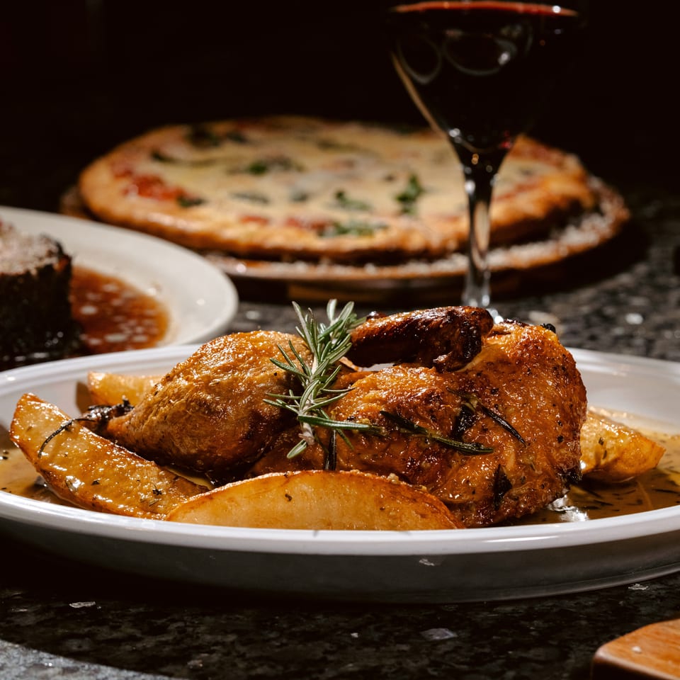Roasted chicken with potatoes and rosemary served on a platter, with steak, pizza, and wine in the background.