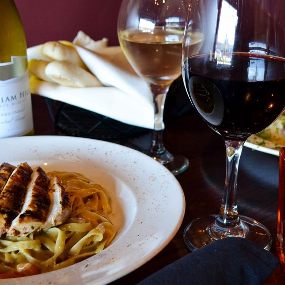 Restaurant table featuring chicken Alfredo pasta, shrimp dish, white and red wine, bread, and a glowing candle.