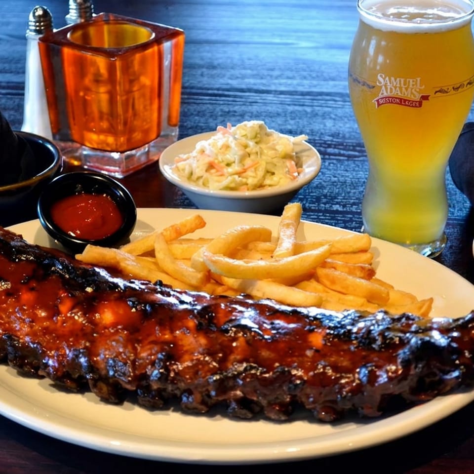 Barbecue-glazed ribs with french fries, coleslaw, dipping sauce, and a Samuel Adams beer on a restaurant table.