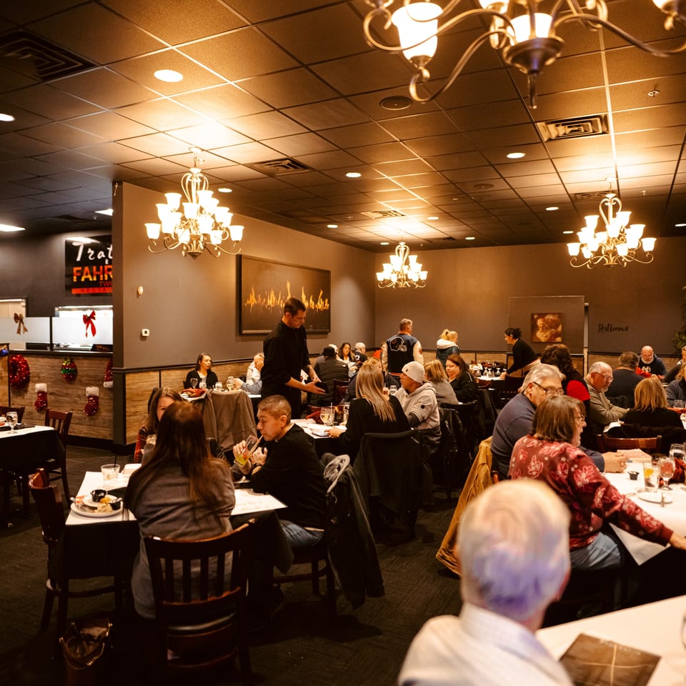 Busy restaurant dining room filled with diners, white-tablecloth tables, chandeliers, festive decorations, and a server attending guests.