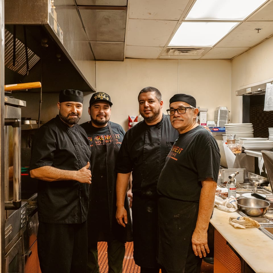 Four restaurant kitchen staff pose together in black uniforms and chef hats amid commercial cooking equipment and stacks of plates.