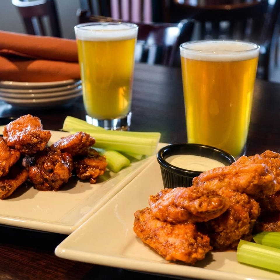 Two plates of saucy chicken wings with celery and dipping sauce, accompanied by two glasses of beer in a restaurant.
