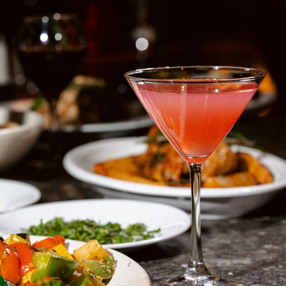 Pink cocktail in a martini glass surrounded by colorful vegetables and plated dishes on a dimly lit restaurant table.