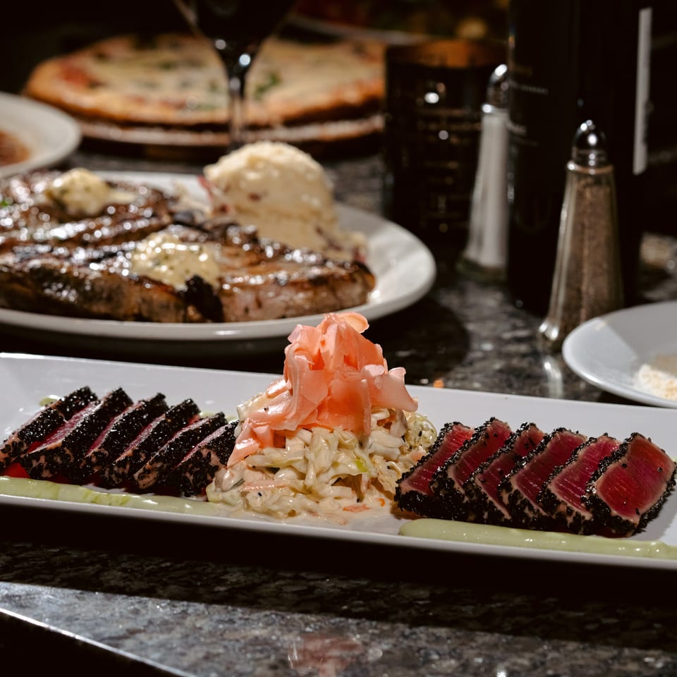 Sliced sesame-crusted seared tuna with coleslaw and pickled ginger, accompanied by steaks and pizzas on a restaurant table.